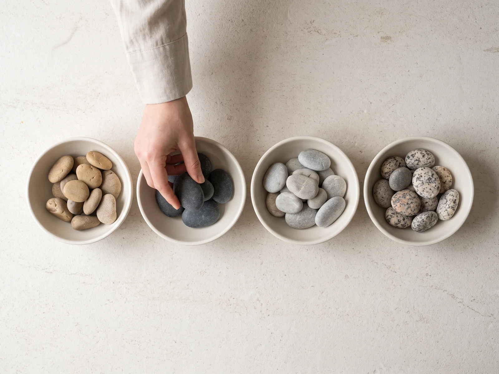 A hand setting a smooth gray pebble into a shallow dish, in a row of dishes of pebbles sorted by tone on a pale limestone table.